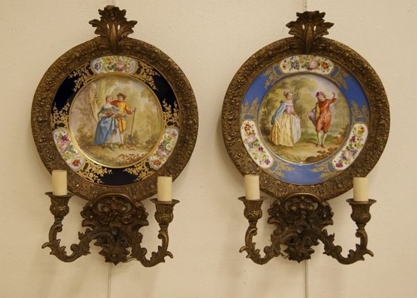 Coppia di appliques in metallo a due luci ciascuno, con piatti in porcellana SevrÃ¨s decorati a scene galanti, l'uno con fondo azzurro l'altro con fondo blu, cm 43x29