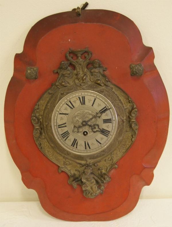 Orologio in metallo dorato sormontato da due  puttini con motivo a coppa, quadrante inciso con motivi vegetali e numeri in nero, supporto in   legno sagomato e dipinto a fondo rosso, sec. XIX