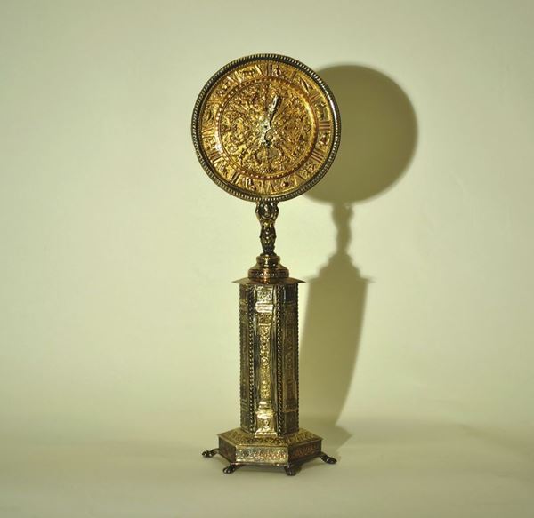Orologio da tavolo, in metallo argentato, quadrante circolare cesellato a  candelabra e segni zodiacali sostenuto da putto poggiante su colonna a sezione esagonale decorata da tralci vegetali, base su piedini a zampa ferina, alt. cm 62, mancanze