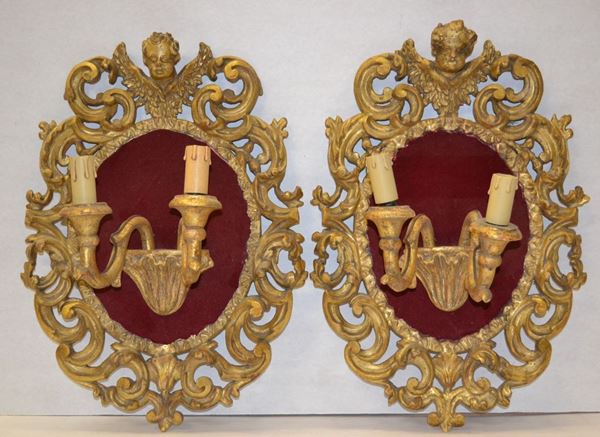 Coppia di appliques, sec. XVIII,  in legno intagliato e dorato a due bracci ciascuna, cornice a volute sormontate da teste di putti, alt. cm 47x34, lievi danni  (2)