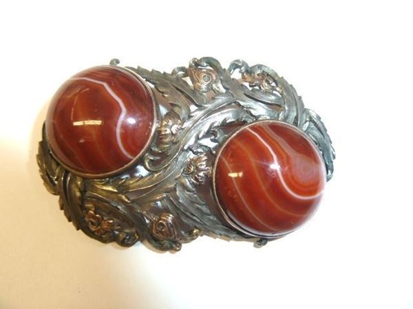 Spilla in argento, oro a basso titolo e corniole                            disegnata come due tralci di fiori e foglie intersecanti fra loro in oro ed argento traforato e cesellato, entro cui sono montate due corniole          circolari cabochon, g 26