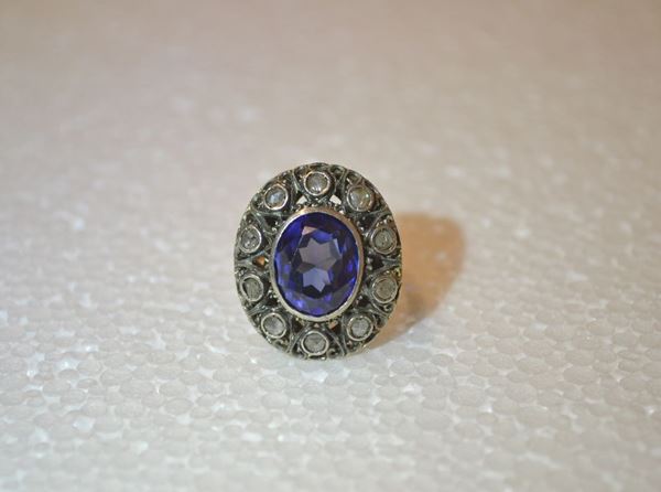 Anello in oro giallo, argento, pietra blu ad imitazione dello zaffiro e     rose di diamante                                                            al centro una pietra ad imitazione dello zaffiro montata entro un castone   traforato e cesellato impreziosito da rose di diamante, g 7