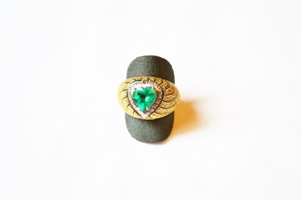 Anello in oro giallo, oro bianco, smeraldo e diamanti, realizzato come una fascia bombata decorata da un motivo ondulato in brillanti e centrata da uno smeraldo di taglio a cuore montato entro castone in oro bianco profilato da piccoli brillanti, g 14