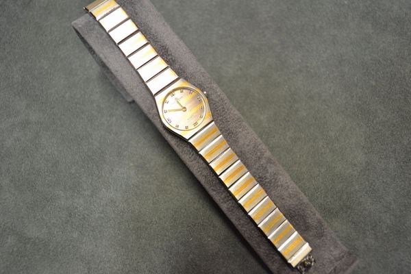 Orologio per signora Baume e Mercier, in acciaio e diamanti Cassa sagomata in acciaio satinato con righe orrizontali dorate, fondello chiuso da quattro viti. Quadrante analogamente decorato con indici in brillanti. Movimento al quarzo. Bracciale a maglie anlogamente decorata alla cassa. Diam. mm 23
