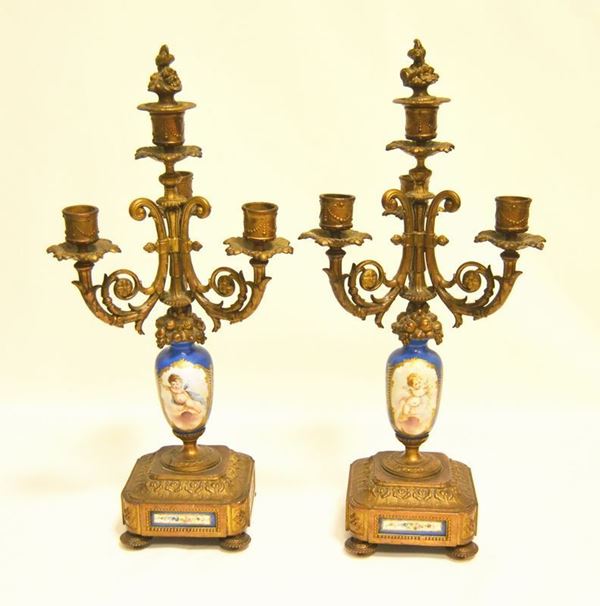 Coppia di candelabri, Francia, sec. XIX, in ottone, a tre luci, bracci lavorati, fusto centrale in ceramica decorato a PUTTI, base quadrata decorata con formella in ceramica, alt. cm 47 ( 2 )