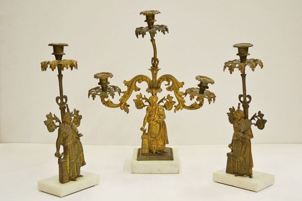 Trittico, fine sec. XIX, composto da una coppia di candelieri ed un         candelabro con fusti raffiguranti personaggi mediorientali, basi in marmo   bianco, alt. cm 37 e cm 47, alcuni danni e mancanze; piccolo orologio,      Francia, fine sec. XIX, in metallo dorato e sbalzato a motivo rocaille,     quadrante in smalto bianco con numeri arabi, alt. cm 21, meccanismo da      revisionare; e piatto commemorativo, in rame, raffigurante al centro Luigi  Augusto Delfino di Francia, tesa sbalazata con girali e palmette, diam. cm  48 (5)