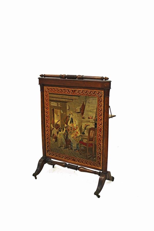 Parascintille, sec. XIX, in mogano, pannello realizzato a piccolo punto raffigurante scena familiare, cimasa a motivo tornito, sostegni mossi desinente in sabot bronzeo a guisa di zampa ferina su rotelle, cm 68,5x44x120
