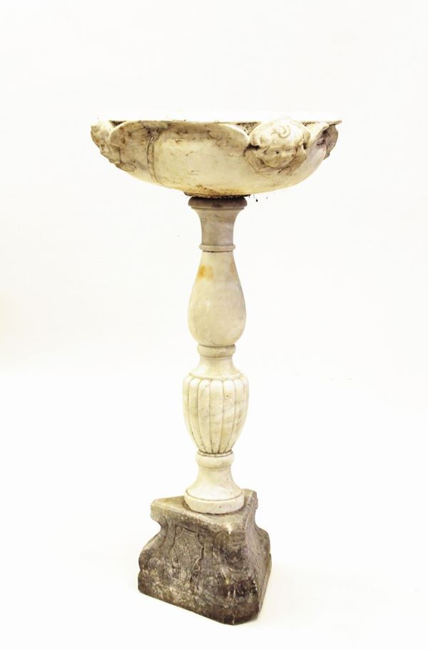 Acquasantiera, sec. XVI, in marmo di Carrara, vasca circolare a orlo everso, impreziosito da quattro rilievi scolpiti a cherubini, fusto ad elementi torniti vasiformi a motivi lisci e baccellati, base a piramide tigonale decorata a stemmi gentilizi, diam. cm 140 alt. cm 75, difetti e restauri