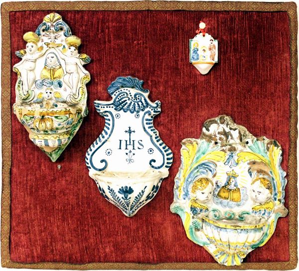 Gruppo di tre piccole acquasantiere in maiolica composte da: una dipinta in policromia con la Madonna di Provenzano affiancata da due cherubini ad altorilievo entro foglie, San Quirico, secolo XVII, alt. cm. 23, vari danni e mancanze; una dipinta in azzurro e bianco con i simboli di Cristo, motivi rocaille nella cimasa e fiori nella vaschetta, Italia centrale, met&agrave; secolo XVIII, alt. cm. 22 e una centrata dalla figura della Madonna di Provenzano dipinta in policromia affiancata da due angeli stanti a bassorilievo, alt. cm. 23, varie mancanze allo smalto nella parte inferiore montate su un pannello rettangolare rivestito in velluto rosso