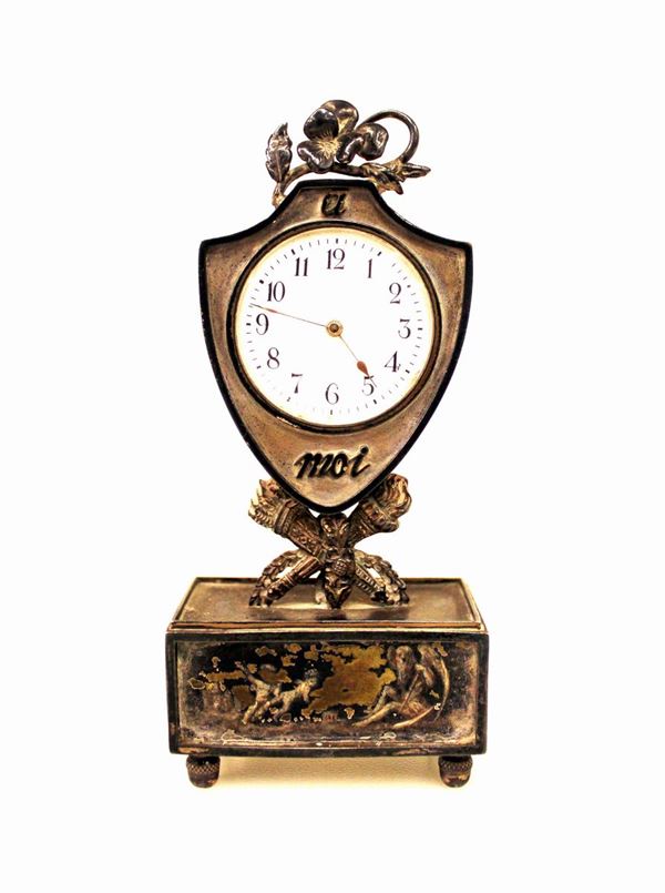 Piccolo orologio da tavolo, fine secolo XIX, in metallo argentato o argento in forma di scudo iscritto a Moi e sormontato da pans&egrave;, poggiante su fiaccole e faretre incrociate, base a parallelepipedo con allegoria del Tempo su piedini a ghianda, alt. cm. 12, pannello frontale con mancanze, completo di chiave