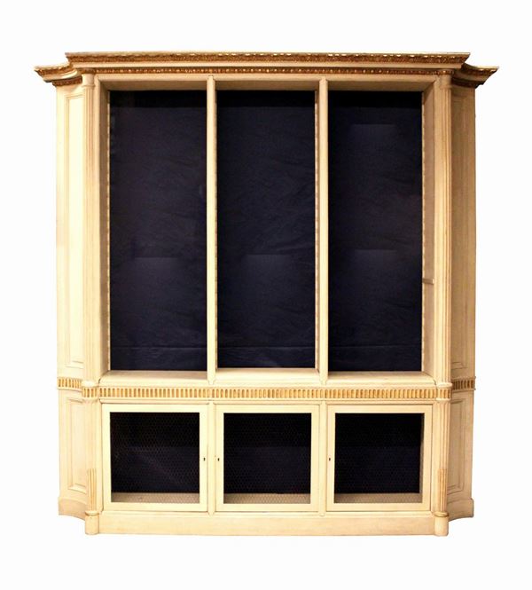 Libreria, in stile neoclassico, in legno laccato e dorato, fronte tripartito, corpo superiore a scaffali regolabili in altezza, corpo inferiore a tre sportelli chiusi da rete metallica, corpi intervallati da tavolette estraibili dal fronte scannellato, fianchi scantonati a lati convessi, colonne rudentate, cimasa modanata a doppio ordine ad ovoli e fogliette, cm 252x54x266, completa di fondi e scaffali, mancante di regoli reggi scaffali, difetti